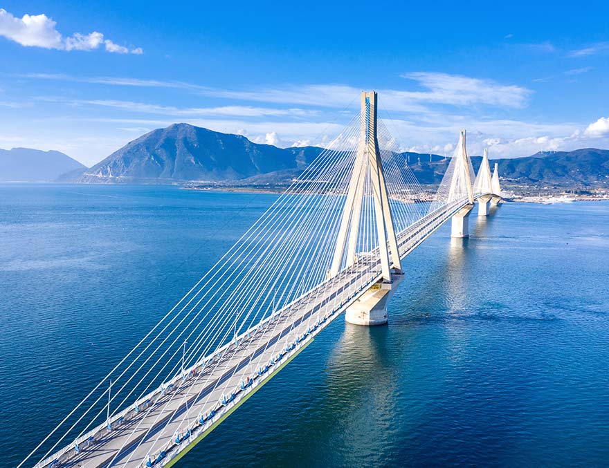 A large bridge over a body of water with a city, mountains and wind turbines in the distance.