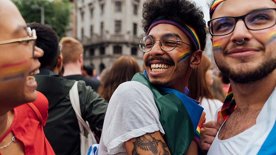 Three people in a crowd with rainbow face paint and rainbow-colored clothing accessories.