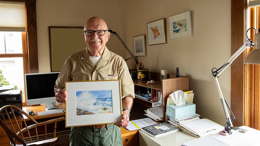 Older adult proudly holding a framed painting in a home office.