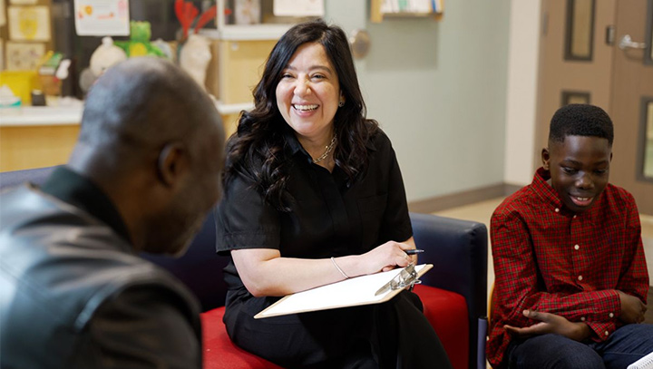 Three people seated in a waiting room; one person holds a clipboard while speaking with an adult and a child.
