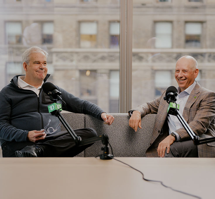 Two people seated on a sofa speaking into microphones during a recorded conversation in an office setting.