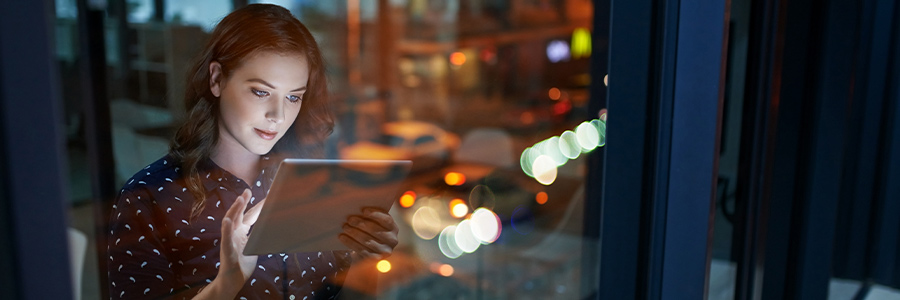 A person using a tablet device near a window at night, with city lights and reflections visible outside