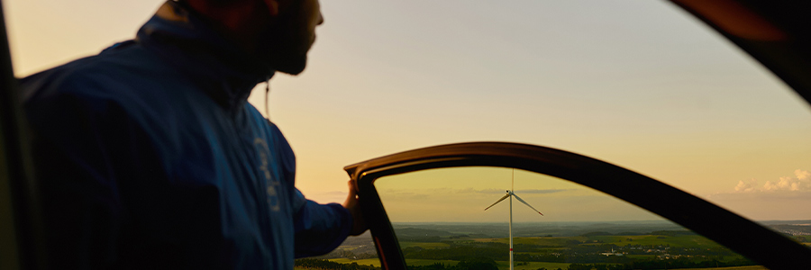 Person looking across a countryside wind farm through an open car door at sunset.