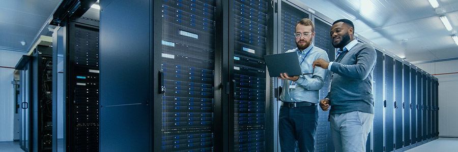 Two people standing in a server room, discussing something on a laptop in front of tall racks of computer servers.