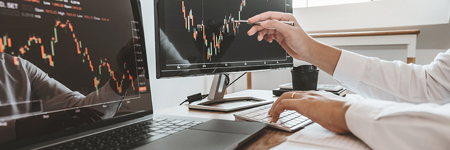 Stock market charts on monitors in a workspace, with a person pointing at one screen.