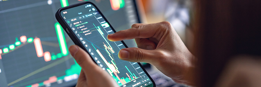 Stock market charts on monitors in a workspace, with a person pointing at one screen.