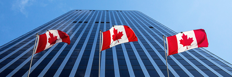 Three Canadian flags in front of a tall glass office building against a blue sky.