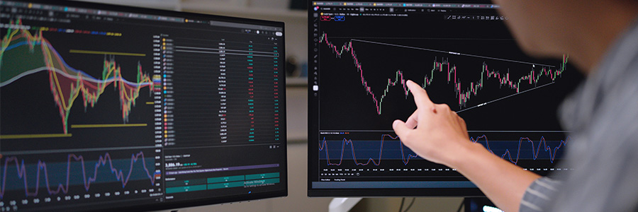 Person pointing at financial charts displayed on dual monitors, showing market trends and technical analysis indicators.