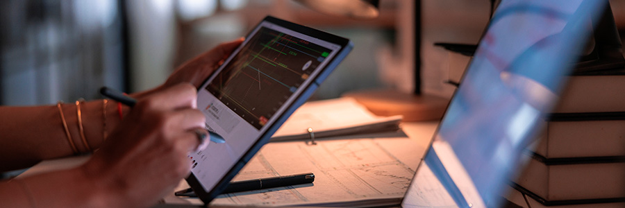 Person uses a stylus on a tablet displaying market charts beside computer screens and papers on a desk.