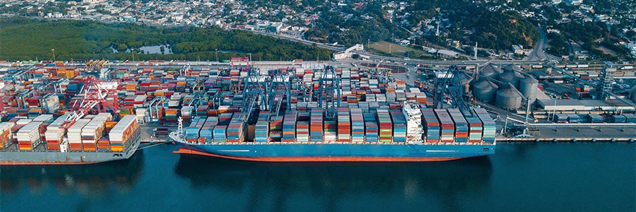 Aerial view of a container ship docked at a busy port with stacked shipping containers and a hillside city in the background.