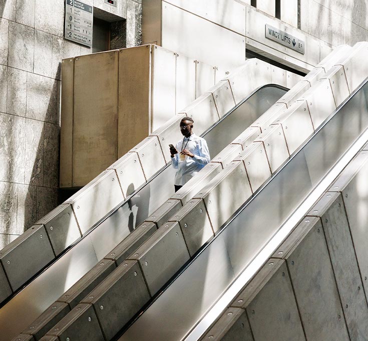 Person using a smartphone while descending a modern indoor escalator with sunlight casting shadows across the structure.