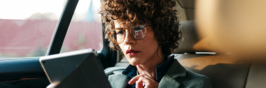 Person sitting in a car’s back seat, using a tablet with one hand and wearing glasses.