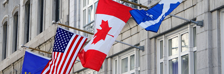 Four flags are displayed on flagpoles attached to the side of a stone building with large windows including the Canadian and American flags. 