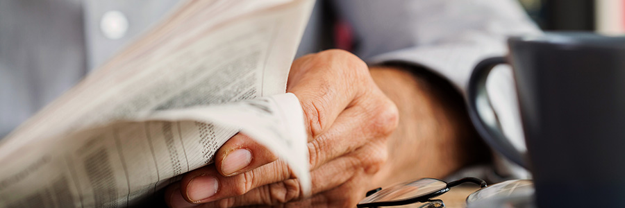 Close-up of hands flipping through a newspaper beside glasses and a coffee mug.