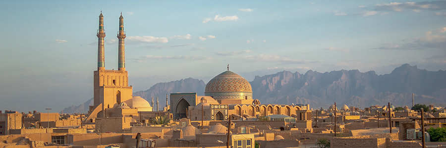 Historic mosque with twin minarets and a tiled dome overlooking a desert city, with mountains in the background.