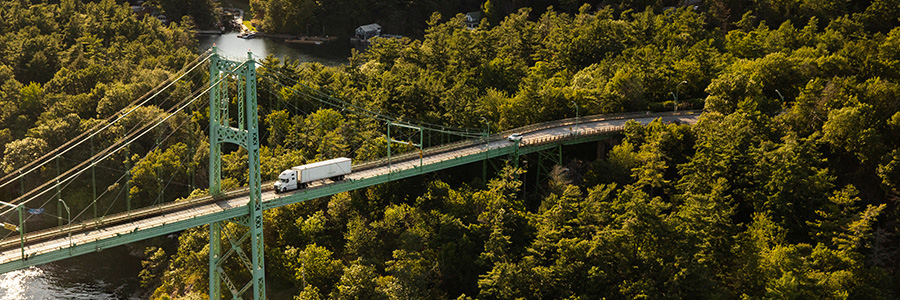 A transport truck crosses a suspension bridge surrounded by dense forest and water.
