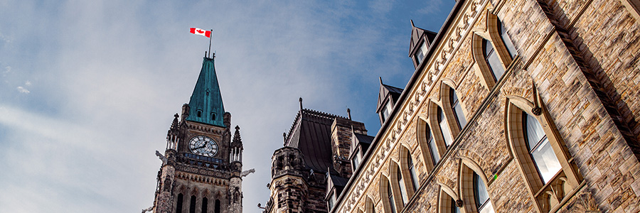 Historic stone Parliament building with the Peace Tower clock and a Canadian flag flying overhead against a partly cloudy sky.