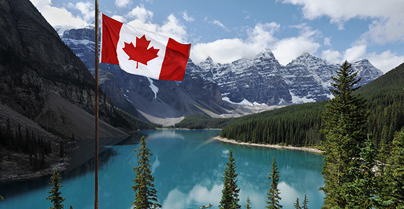 A Canadian flag waves in the forefront of a mountain landscape