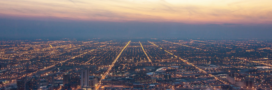 Vue aérienne d’une ville au crépuscule, où les rues illuminées s’étendent jusqu’à l’horizon sous un ciel aux couleurs éclatantes du coucher du soleil.