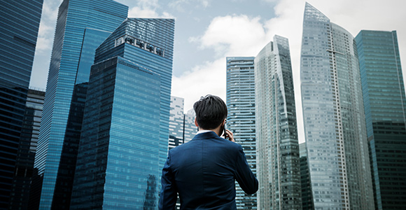 A man stands in front of multiple city skyscrapers while holding a cellphone to his ear.