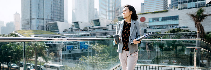 Business professional walking on an urban terrace with a coffee and documents, city skyline in the background.
