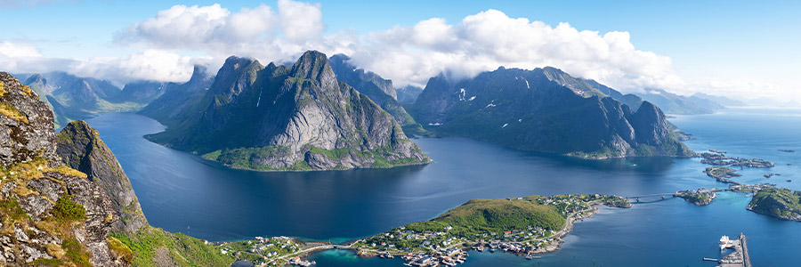 Wide landscape view of a coastal fjord with steep mountains, blue water and a small seaside village below.