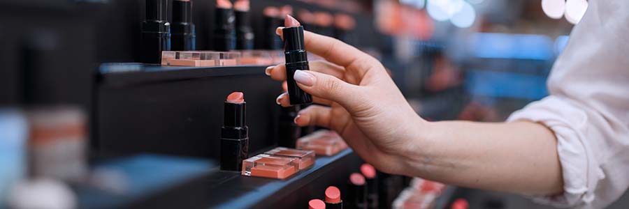 Close up of a person’s hand selecting a lipstick from a display shelf filled with various shades of lipsticks in a cosmetics store.