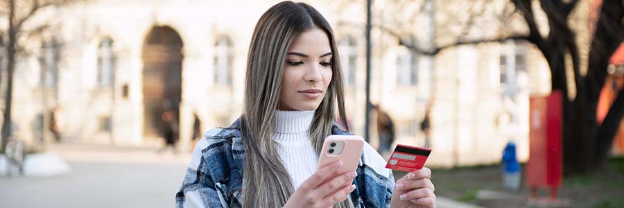 A young person standing outdoors holding a smartphone in one hand and a red credit card in the other.