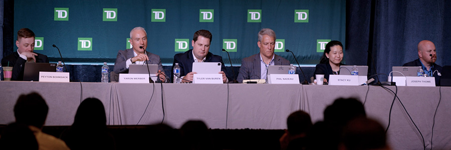 Several people sitting behind a table in front of an audience as part of a panel of speakers. The wall behind them is adorned with the TD logo.