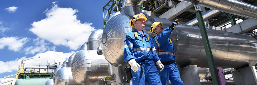 Industrial workers inspecting large metal pipes at an energy facility under a blue sky.