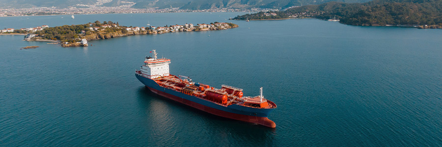 Tanker ship sailing in a blue coastal bay with small islands and mountains in the background.