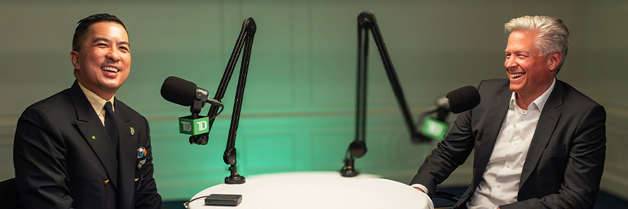 Two speakers seated at a podcast table with microphones, recording a conversation in a studio setting.