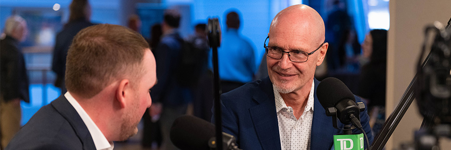 Kyle Boucher (left) and Kim Kelderman (right) in conversation at a podcast-style setup with microphones in a busy event space.