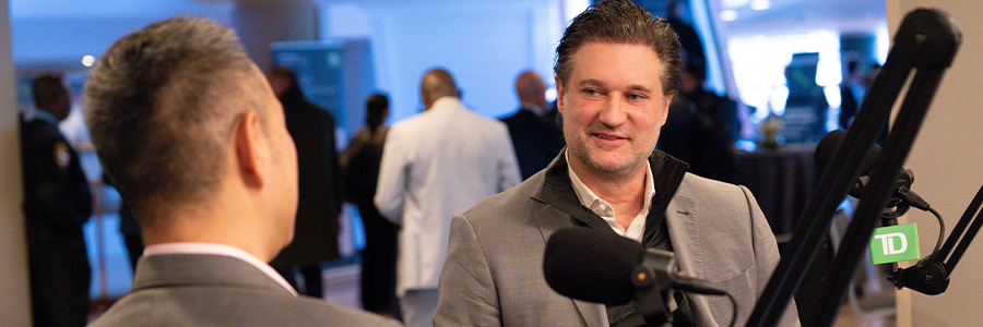 Podcast interview setup at a healthcare conference, with a Charles Rhyee and Randall Stanicky speaking into microphones in a busy event space.