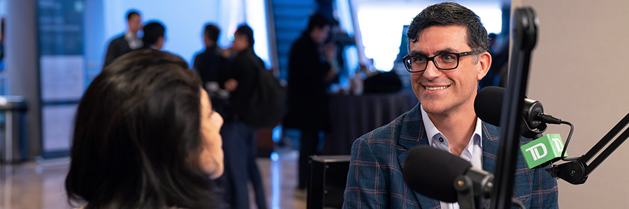 Ritu Baral and Marcio Souza are seated at microphones recording a podcast at a conference venue. 