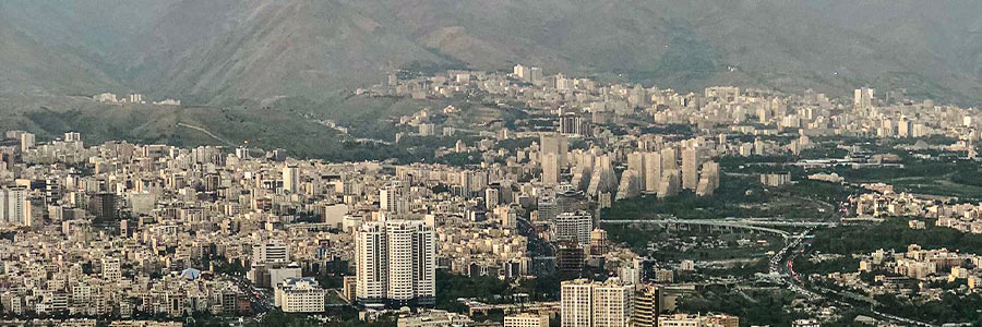 Aerial view of a dense city skyline with high-rise buildings, highways, and green spaces set against a mountainous backdrop.