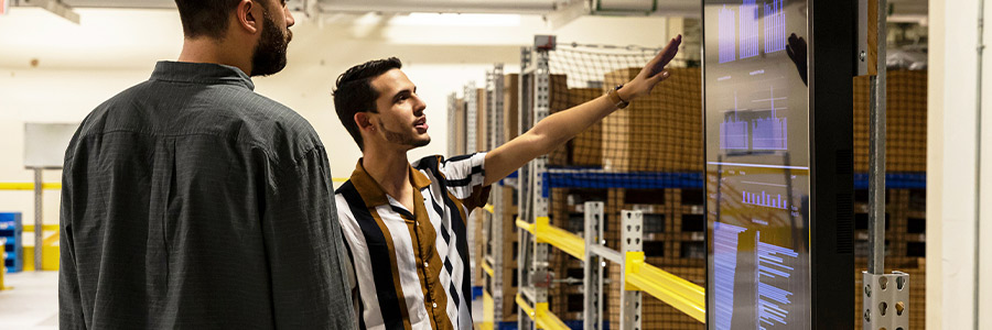 Warehouse staff reviewing performance data on a large digital dashboard beside storage racks.