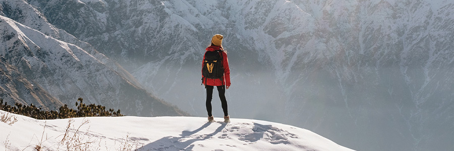Person in a red winter jacket with a backpack standing on a snowy mountain ridge overlooking snow-covered peaks.