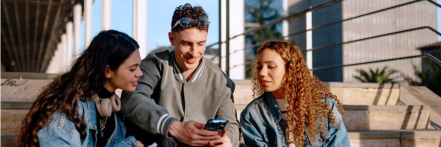 Three young college-age students sitting on outdoor steps, talking while holding notebooks and mobile phone.