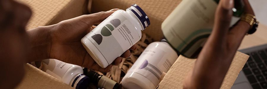 Person holding supplement bottles above an open shipping box filled with health products.