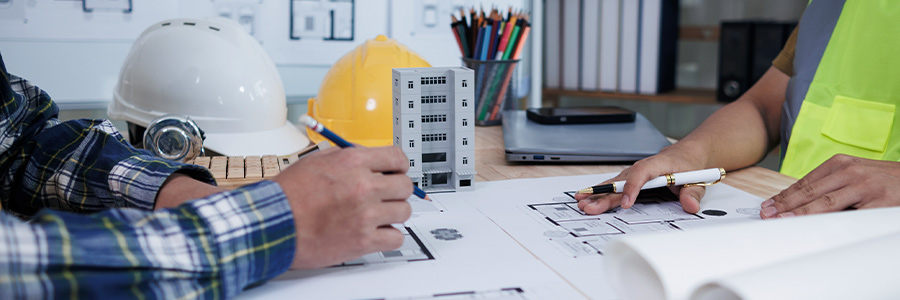 Two people reviewing architectural blueprints at a desk with hard hats and a building model.