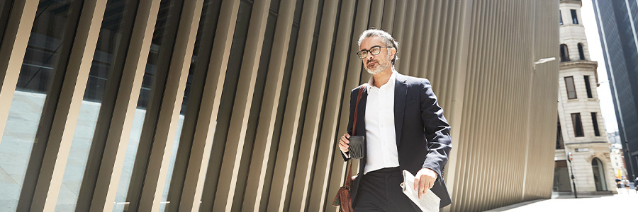 Person in business attire walking beside a modern building, carrying a mug and documents on a city street.