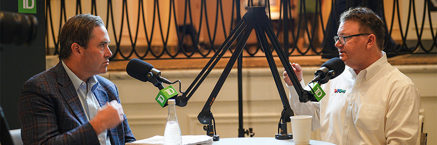 Two participants record a podcast interview at a round table with TD-branded microphones, papers and drinks in a warmly lit indoor setting.