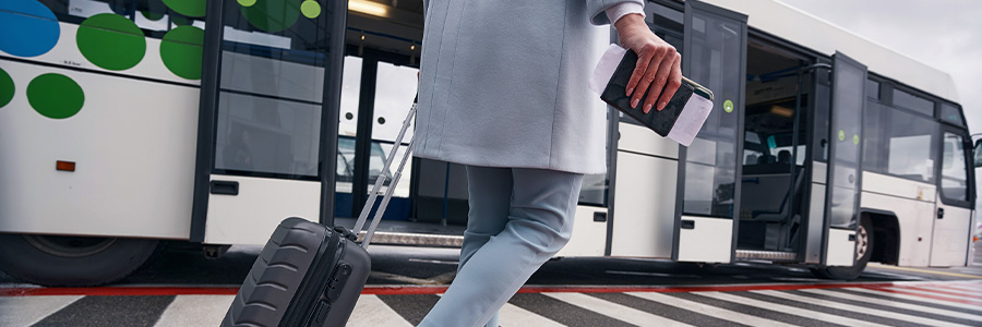 Person walking with a suitcase towards an airport shuttle bus, holding travel documents.