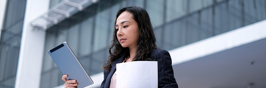 A person outside a modern office building holding a tablet computer and documents.