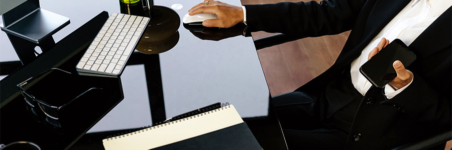 Professional seated at desk using computer mouse, with smartphone, notebook and keyboard on workspace.