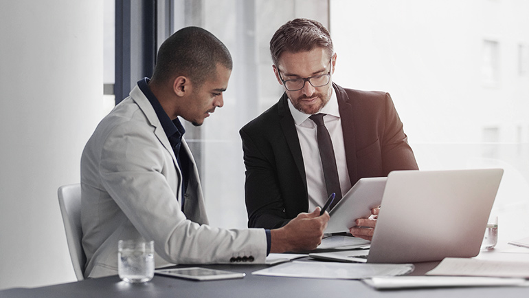 Two men sitting in front of a laptop looking at reports. 