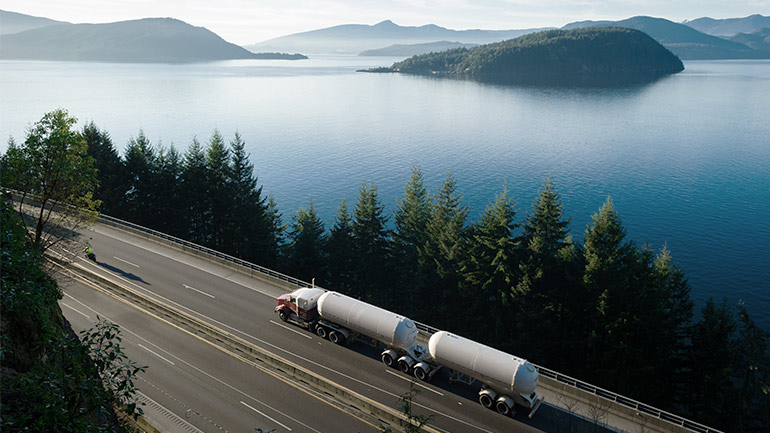 A hydrogen transport vehicle drives on a highway beside a scenic view of lakes and hills.