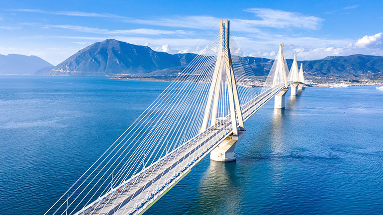 A large bridge over a body of water with a city, mountains and wind turbines in the distance.