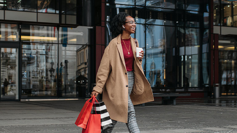Person walking outside a modern office building holding shopping bags and a takeaway coffee cup.